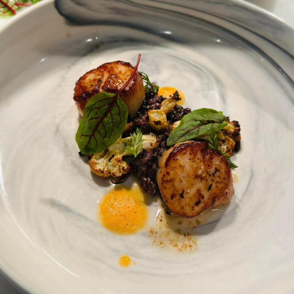 Two seared scallops with black rice, roasted cauliflower, and orange foam on a white plate.