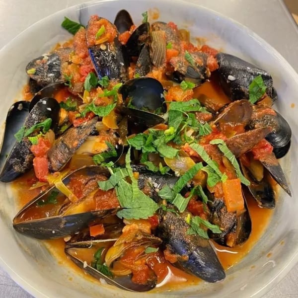 Steamed mussels in a rich tomato broth, garnished with fresh herbs, served in a white bowl.