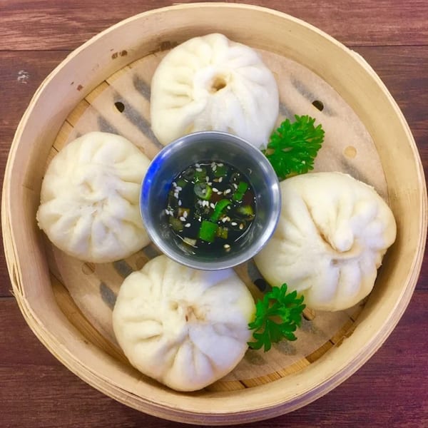 Four steamed buns (baozi) with a dipping sauce in a bamboo steamer, garnished with parsley.