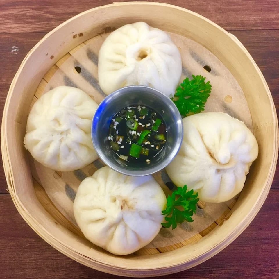 Four steamed buns (baozi) with a dipping sauce in a bamboo steamer, garnished with parsley.